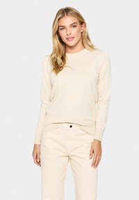 Svetle bež pleteni pulover s krožnim izrezom, rebrastimi manšetami in robom. Ujemajoč se z svetlimi hlačami in nevtralno ozadje.