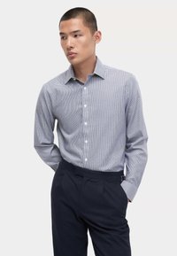 Camicia button-down a scacchi blu e bianchi, maniche lunghe, colletto, abbinata a pantaloni blu navy, con una texture liscia e un design aderente.