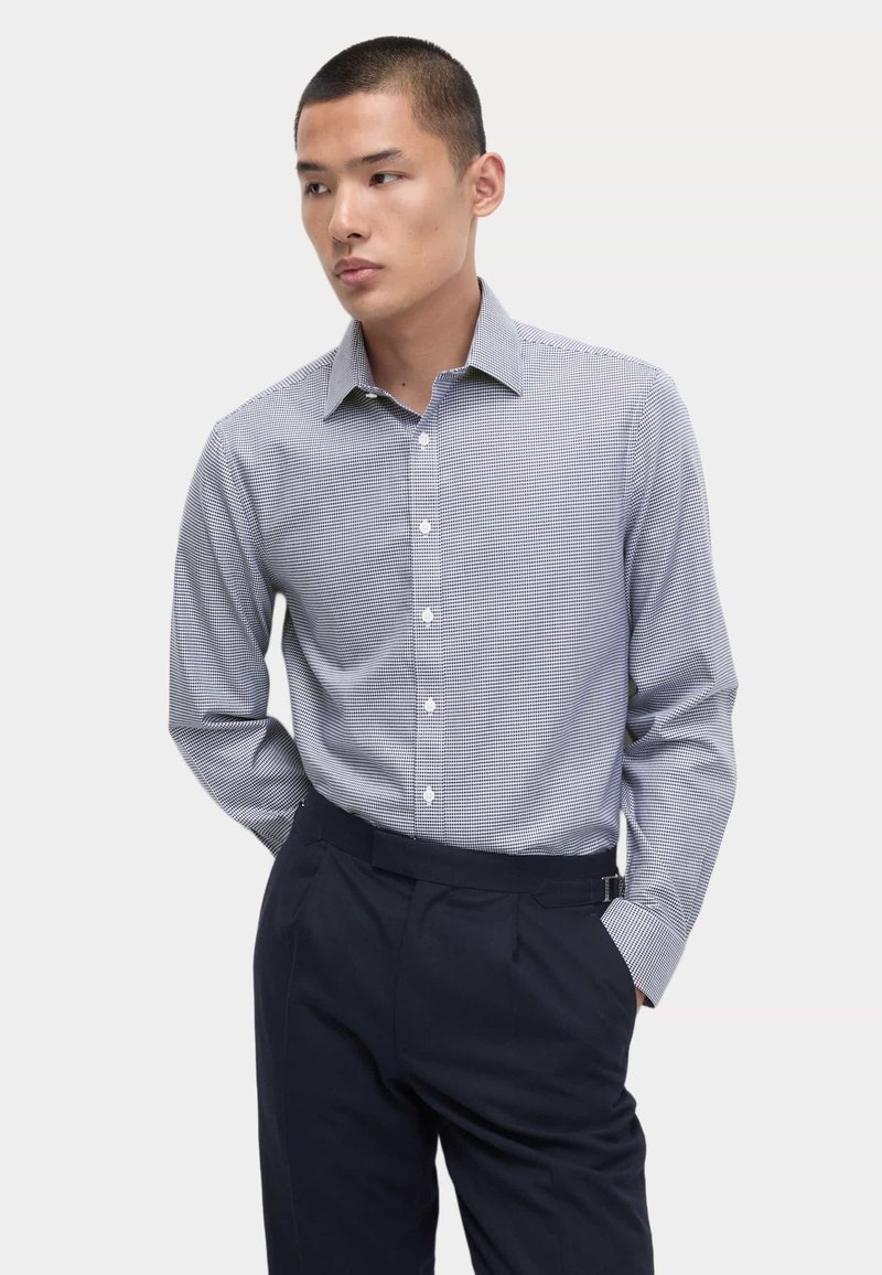 Camicia button-down a scacchi blu e bianchi, maniche lunghe, colletto, abbinata a pantaloni blu navy, con una texture liscia e un design aderente.