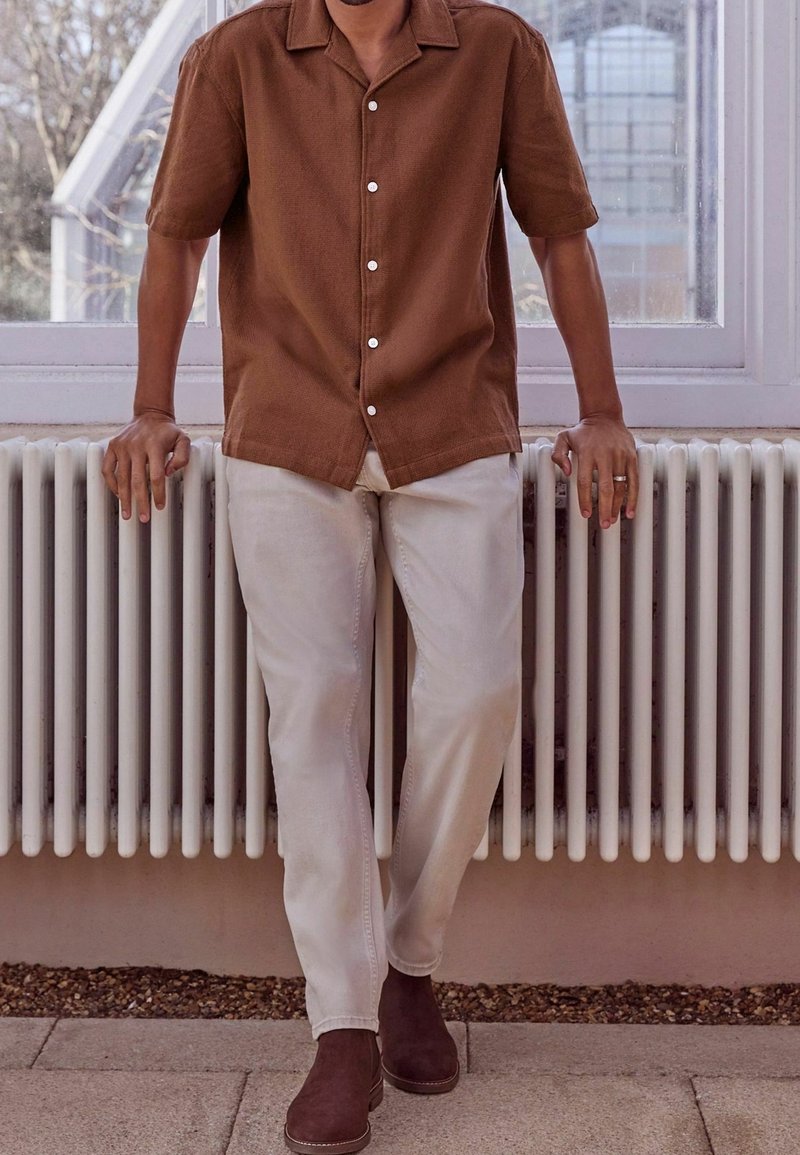 Brązowa koszula z krótkim rękawem o fakturowanej powierzchni, białe guziki, zestawiona z jasnokremowymi spodniami i ciemnobrązowymi botkami za kostkę. W tle znajduje się grzejnik.