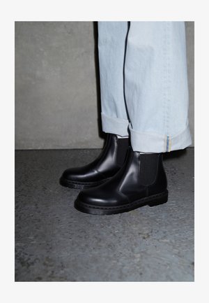 Botas Chelsea pretas de pele com painéis laterais elásticos e sola de borracha texturizada, combinadas com jeans de ganga azul clara com bainha dobrada.