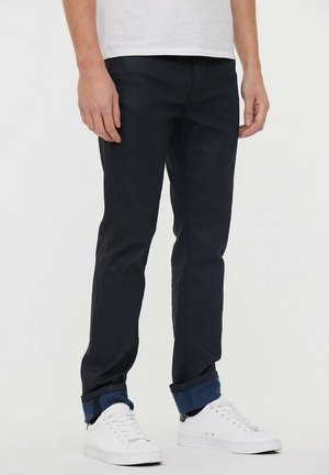 Jean slim-fit bleu foncé en tissu résistant avec une texture lisse, présentant des ourlets roulés et un design classique à cinq poches.