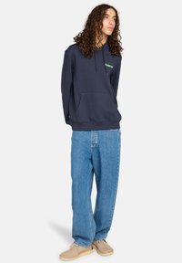 Felpa navy in tessuto morbido con tasca frontale, cappuccio con cordino e logo verde. Abbinata a jeans baggy azzurri chiari e scarpe beige.