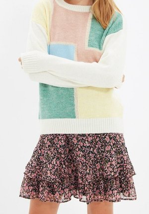 Femme portant un pull pastel à blocs de couleurs et une mini-jupe noire fleurie à volants, bras croisés.