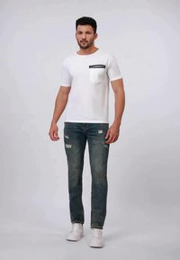 Hombre de pie vestido con una camiseta blanca con bolsillo en el pecho, jeans azules desgastados y zapatillas blancas, frente a un fondo liso.