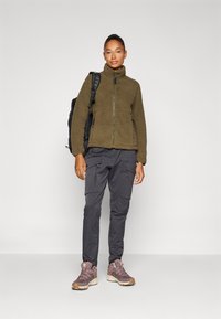 Olivenfarget fleece-jakke med glidelås foran, kombinert med mørke cargo-bukser og lilla tursko, som har en teksturert overflate, samt en ryggsekk.