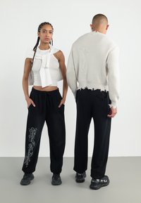 White cropped top with bow detail, paired with black sweatpants featuring white script. Sweater is light gray with frayed hems. Both wear sneakers.