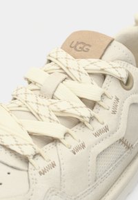 Baskets beige clair avec dessus en daim texturé, lacets crème contrastants ornés d'un motif en losange, et patch logo UGG marron sur la languette.