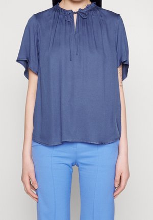 Kobieta ubrana w luźną, ciemnoniebieską bluzkę z krótkim rękawem, z wiązanym dekoltem w kształcie dziurki od klucza i jasnoniebieskie spodnie.