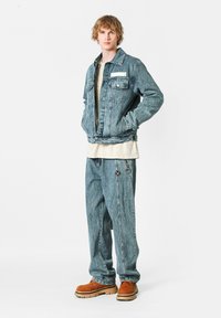 Chaqueta de mezclilla con un acabado azul claro, dos bolsillos y una etiqueta blanca; combinada con pantalones de mezclilla azul claro y zapatos marrones con una suela gruesa.