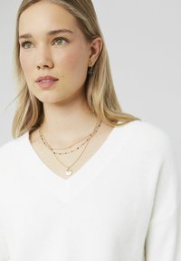 Pull blanc à col en V ; colliers en or superposés avec des détails circulaires et perlés ; le modèle porte de petites boucles d'oreilles rondes avec un design à motifs.