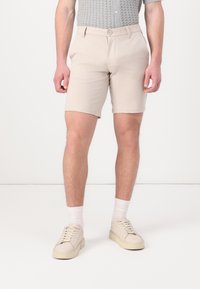 Shorts bej dintr-un material cu o textură netedă, având închidere cu nasture, buzunare laterale și o croială adaptată, asortați cu încălțăminte bej.