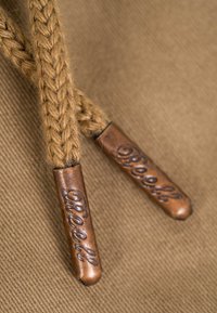 Extremos trenzados de cordón de color beige con puntas de metal color cobre grabadas con "Bleeck" sobre un fondo de tela a juego.