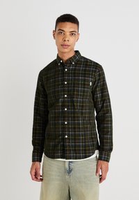 Dark green plaid button-up shirt made of soft fabric, featuring white buttons, a chest pocket, and a curved hem. Worn over light jeans.