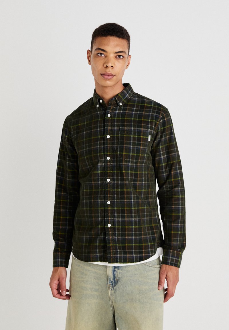 Camisa de cuadros verde oscuro de botones hecha de una tela suave, con botones blancos, un bolsillo en el pecho y un dobladillo curvado. Llevada sobre jeans claros.