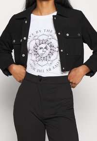 Chaqueta de mezclilla negra con bolsillos frontales y detalles en plata, usada sobre una camiseta blanca con un gráfico del sol y la luna, combinada con pantalones negros.