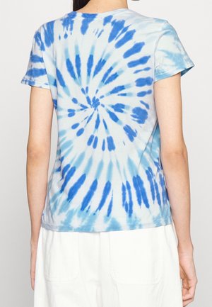 Kobieta ubrana w niebiesko-biały t-shirt tie-dye z krótkim rękawem, pokazana od tyłu, w połączeniu z białymi spodniami lub spódnicą.