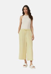 Lichtgele wijde broek met een elastische tailleband en trekkoord; gecombineerd met een aansluitend beige ribbel tanktop en witte sandalen.