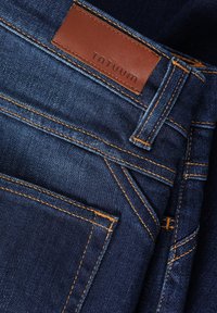 Tmavomodré džíny z denimu s hnedým koženým štítkom na páse s nápisom "TNTUUM", s kontrastným oranžovým prešívaním a klasickým dizajnom so štyrmi vreckami.