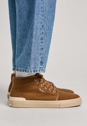 Zapatillas altas de ante marrón con suelas beige, que cuentan con cordones redondos y un logo en el lateral. Llevadas con jeans de corte recto azul claro.