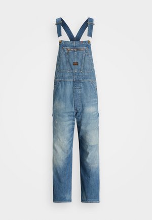 Salopete din denim, cu bretele albastre reglabile, un buzunar frontal și o nuanță de albastru decolorat, având detalii cu petice pe picioare.