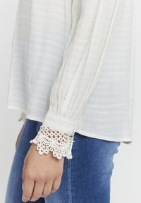 Weiße Bluse mit langen Ärmeln aus strukturiertem Stoff und Spitzenmanschetten, kombiniert mit blauen Jeans, die ein entspanntes, lässiges Outfit-Detail zeigen.