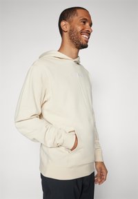 Kremowy hoodie wykonany z miękkiego materiału, z przednią kieszenią i sznurkiem w kapturze. Dyskretny haftowany logo na piersi.