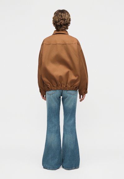 Άτομο όρθιο με την πλάτη προς τον θεατή, φοράει φαρδιά καφέ σακάκι και μπλε τζιν παντελόνι με φαρδιά πόδια, σε απλό φωτεινό φόντο.