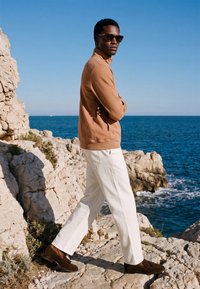 Homme portant des lunettes de soleil, une veste marron, un pantalon blanc et des mocassins foncés, marchant sur des falaises rocheuses au bord de l'océan sous un ciel bleu clair.