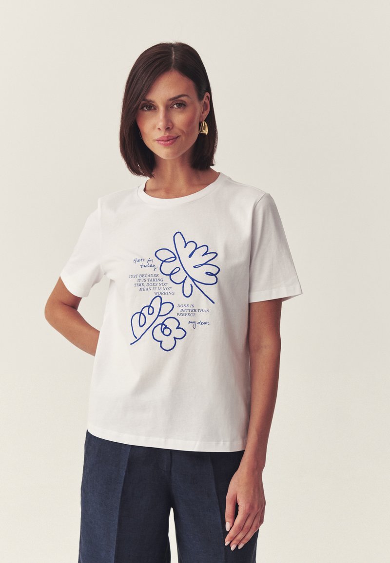 Femme aux cheveux courts et foncés portant des boucles d'oreilles en or et un t-shirt blanc avec un motif floral bleu en ligne et un texte motivant, debout devant un fond uni.