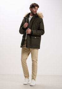 Uomo che indossa un parka verde oliva con cappuccio in pelliccia, pantaloni beige, sneakers bianche e maglione bianco su sfondo bianco.