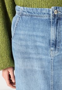 Világoskék farmer rövidnadrág elülső zsebekkel, enyhe koptatással és fém gombos záródással. Zöld kötött pulóverrel párosítva.