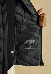 Čierna prešívaná bunda so syntetickým polstrovaním, zapínaním na zips a označením značky Joules. Obsahuje postrannú vrecko na úložný priestor.