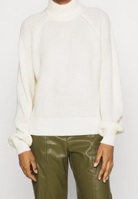 Maglione bianco spesso lavorato a maglia con un colletto alto, polsini a coste e vestibilità comoda, abbinato a pantaloni aderenti verde oliva.