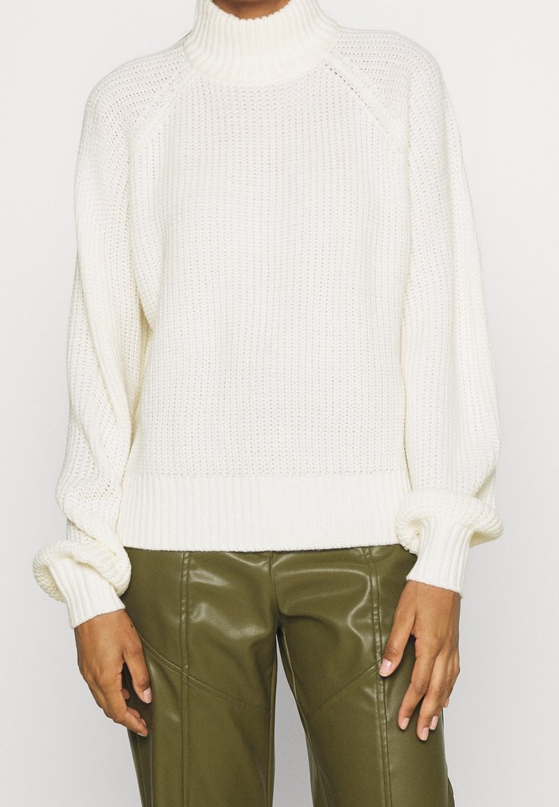 Maglione bianco spesso lavorato a maglia con un colletto alto, polsini a coste e vestibilità comoda, abbinato a pantaloni aderenti verde oliva.