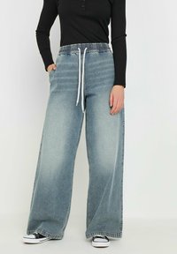 Pantaloni in denim a gamba larga di colore azzurro chiaro con vita elastica e cordoncino bianco. Abbinati a una maglia nera a maniche lunghe e sneakers nere.