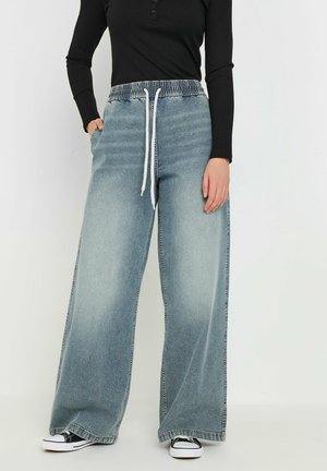 Lichtblauwe denim broek met wijde pijpen, voorzien van een elastische tailleband en witte trekkoord. Gestyled met een zwart langemouwen shirt en zwarte sneakers.