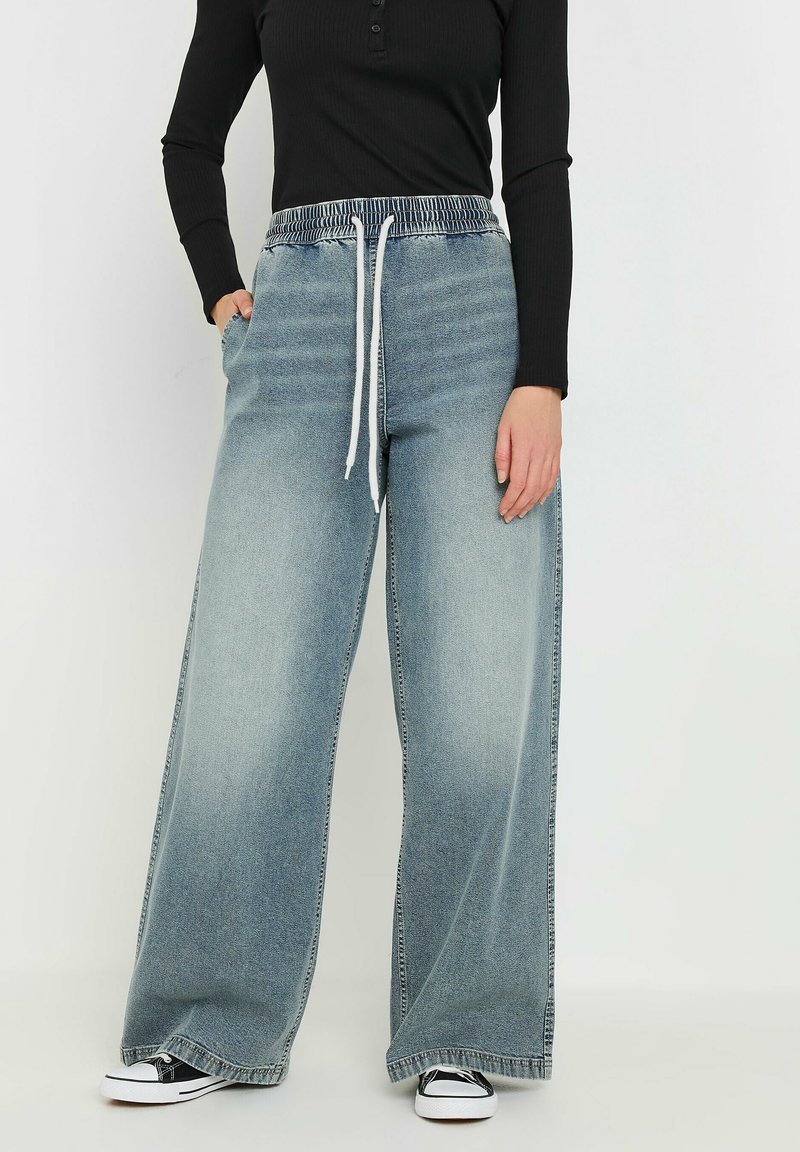 Pantaloni in denim a gamba larga di colore azzurro chiaro con vita elastica e cordoncino bianco. Abbinati a una maglia nera a maniche lunghe e sneakers nere.