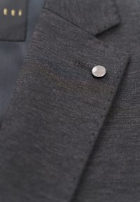 Dunkelgrauer Jackenstoff mit subtiler Struktur, versehen mit einem metallischen Knopf und einem kleinen Etikett, das maßgeschneiderte Details und klare Linien zeigt.