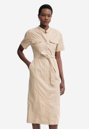 Donna che indossa un vestito midi beige con bottoni, maniche corte, tasche sul petto e cintura in vita, in piedi davanti a uno sfondo bianco.