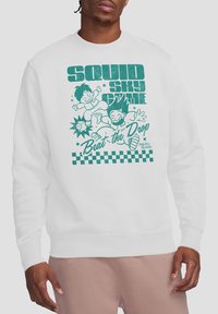 Man wearing a white sweatshirt with a colorful illustration featuring two cartoon characters and the text “Beat the Drop.”