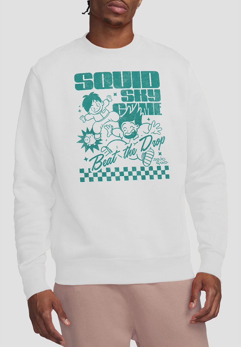 Man wearing a white sweatshirt with a colorful illustration featuring two cartoon characters and the text “Beat the Drop.”