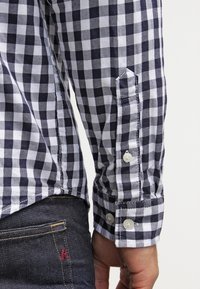 Camicia a quadri in cotone blu navy e bianco, con polsini abbottonati, cuciture in contrasto e una vestibilità casual. Jeans in denim scuri sono parzialmente visibili.
