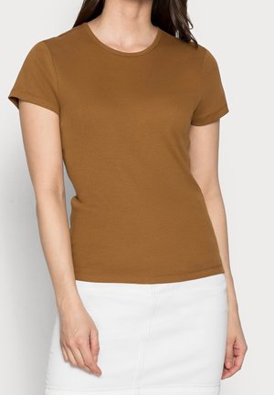 Kobieta ubrana w brązowy dopasowany T-shirt z krótkim rękawem i białą spódnicę na jednolitym jasnym tle.