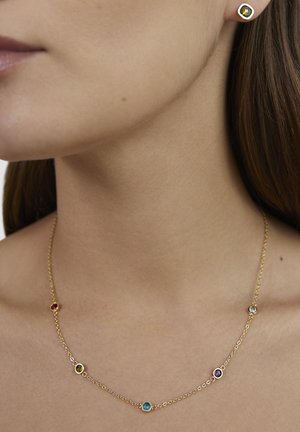 Gouden ketting met kleine, meerkleurige edelstenen gedragen om de hals, gecombineerd met een gouden oorknopje met een gefacetteerde gele steen.