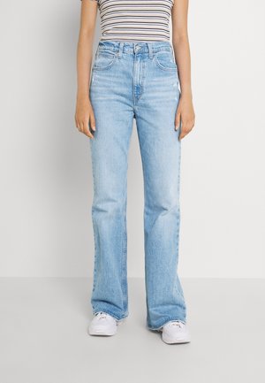 Personne portant un jean bleu clair taille haute et coupe droite, un haut sans manches rayé et des baskets blanches, debout devant un fond uni.