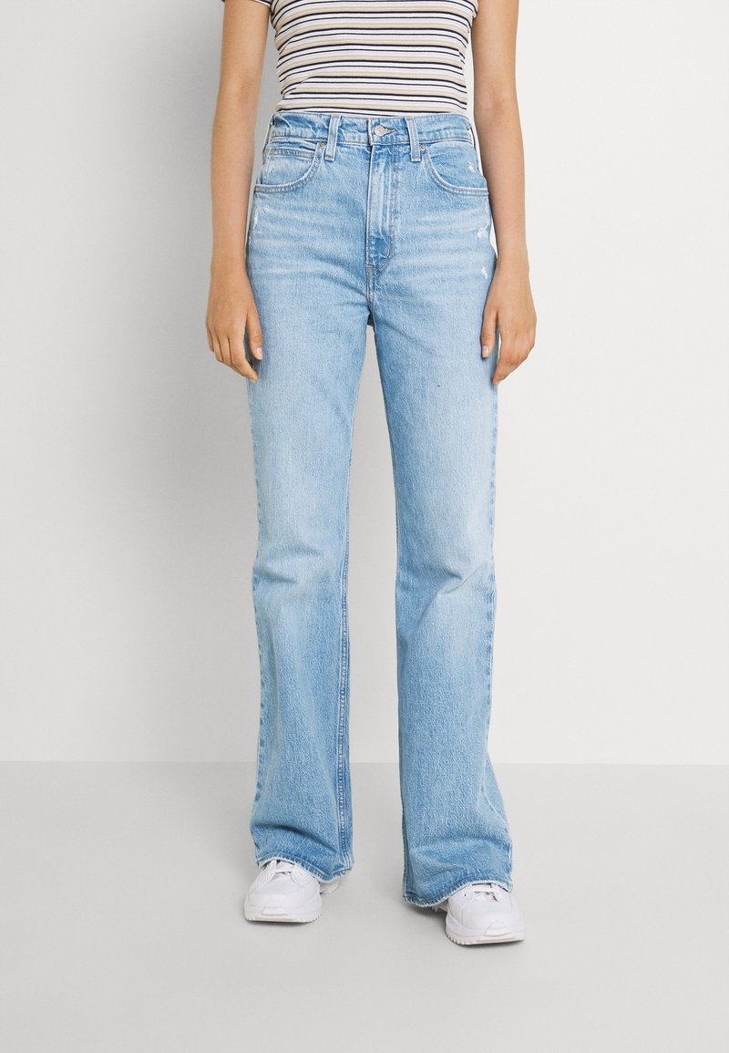 Personne portant un jean bleu clair taille haute et coupe droite, un haut sans manches rayé et des baskets blanches, debout devant un fond uni.