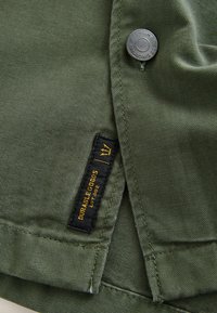 Olivgrüner Stoff mit einer strukturierten Webart, versehen mit einem runden grauen Metallknopf und einem schwarzen Etikett mit goldener Schrift, auf dem steht "DURABLE GOODS Lot 082."