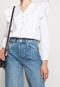 White button-up blouse with ruffle sleeves, textured accents along the front, paired with high-waisted, light blue denim jeans.