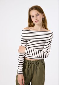 Schulterfreies, geripptes Oberteil in Weiß mit schwarzen horizontalen Streifen. Kombiniert mit locker sitzenden olivgrünen Hosen mit einem elastischen Bund.